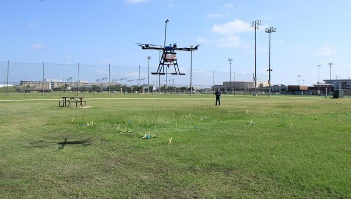 The image shows a remotely piloted aerial application system (RPAAS) drone hovering over a grass field with a person standing nearby. The drone is being used for a weed control application test in an open space.