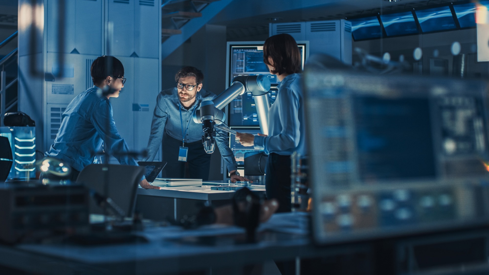 Team of Engineers with Laptop and a Tablet Analyse and Discuss How a Futuristic Robotic Arm Works and Moves a Metal Object.