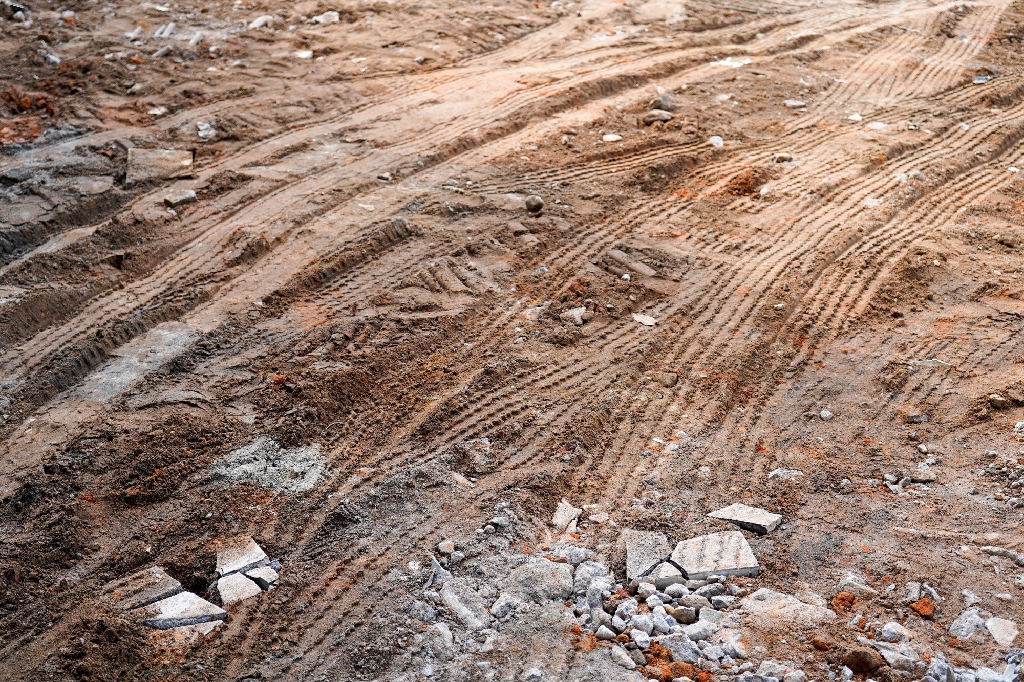 Image shows tire tracks on a brown dirt ground at a construction site. The ground is uneven and shows signs of heavy machinery movement.